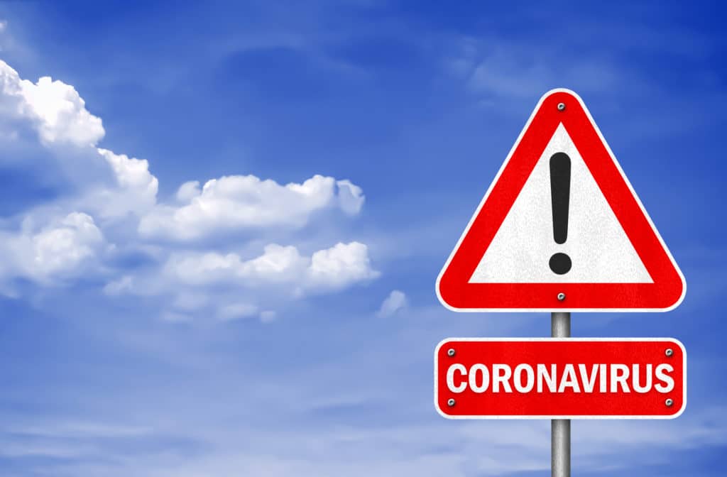 A red warning sign with an exclamation mark and the word "CORONAVIRUS" against a blue sky with clouds.