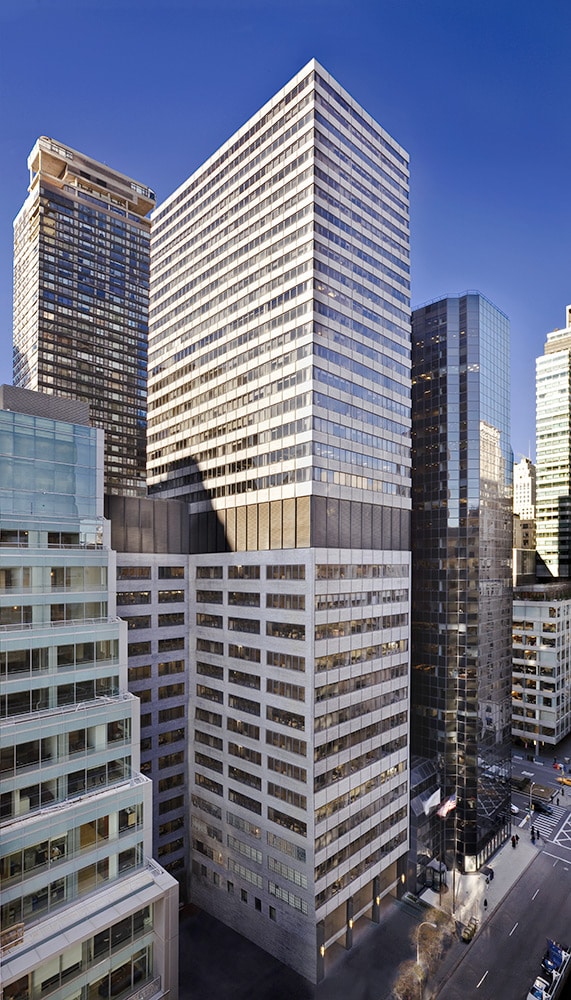 A tall office building with reflective glass windows stands among other high-rise structures on a clear day in a downtown area. An intersection is visible in the lower right corner.