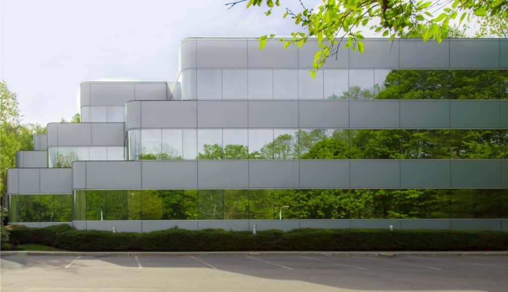 Modern office building with reflective glass facade, surrounded by trees. The building is multi-storied with a tiered design. The parking lot in front is empty.