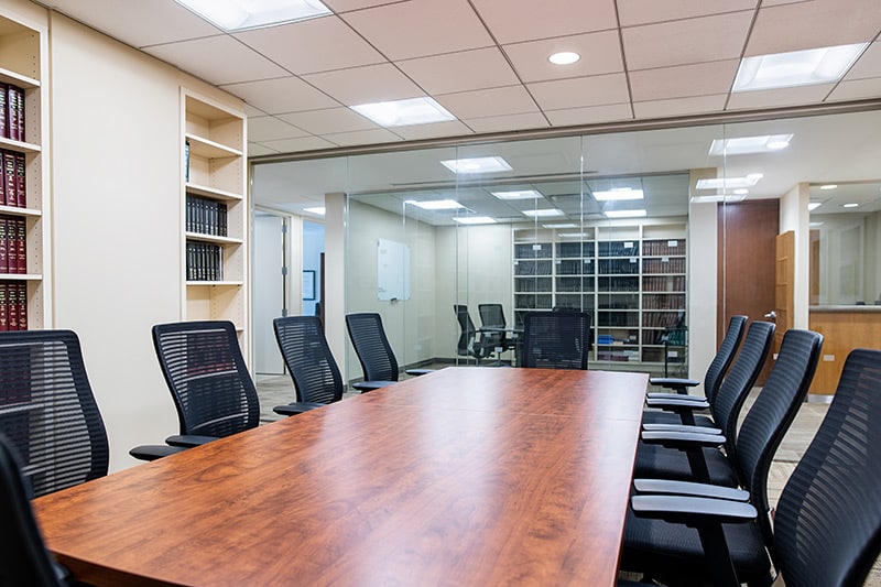 A conference room with a wooden table, black mesh chairs, and glass walls, surrounded by bookshelves and office equipment.