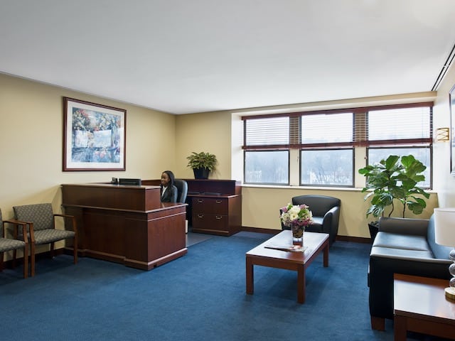 A woman seated at a reception desk in an office waiting area with chairs, a coffee table, a couch, and windows.