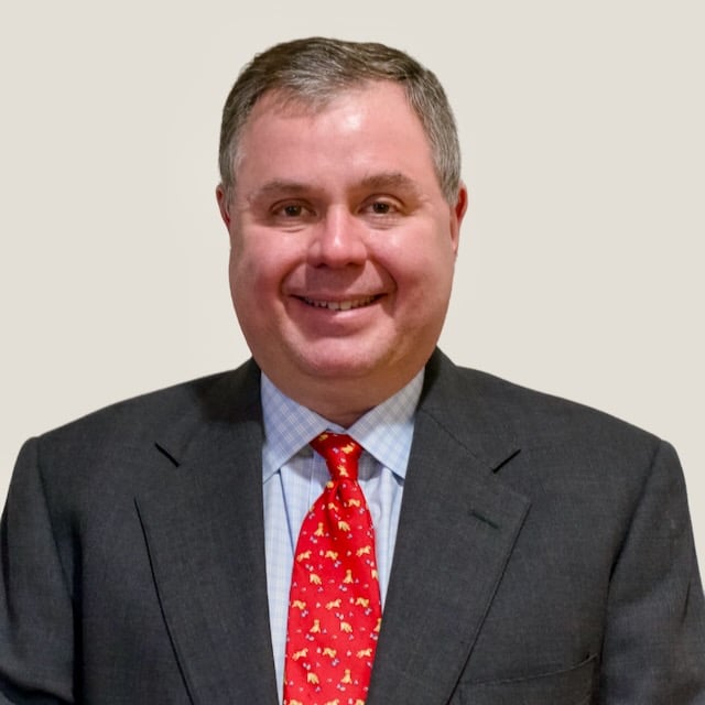 A person wearing a dark suit, light blue checkered shirt, and red patterned tie, smiling against a plain background.