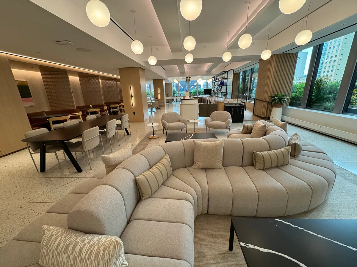 Modern lounge area with a large curved sectional sofa, cushioned dining chairs, round pendant lights, and large windows providing a view of buildings outside.