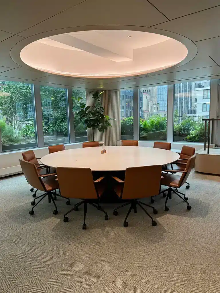 A modern meeting room with a large round table surrounded by ten brown chairs, large windows offering a cityscape view, and a central ceiling light fixture.