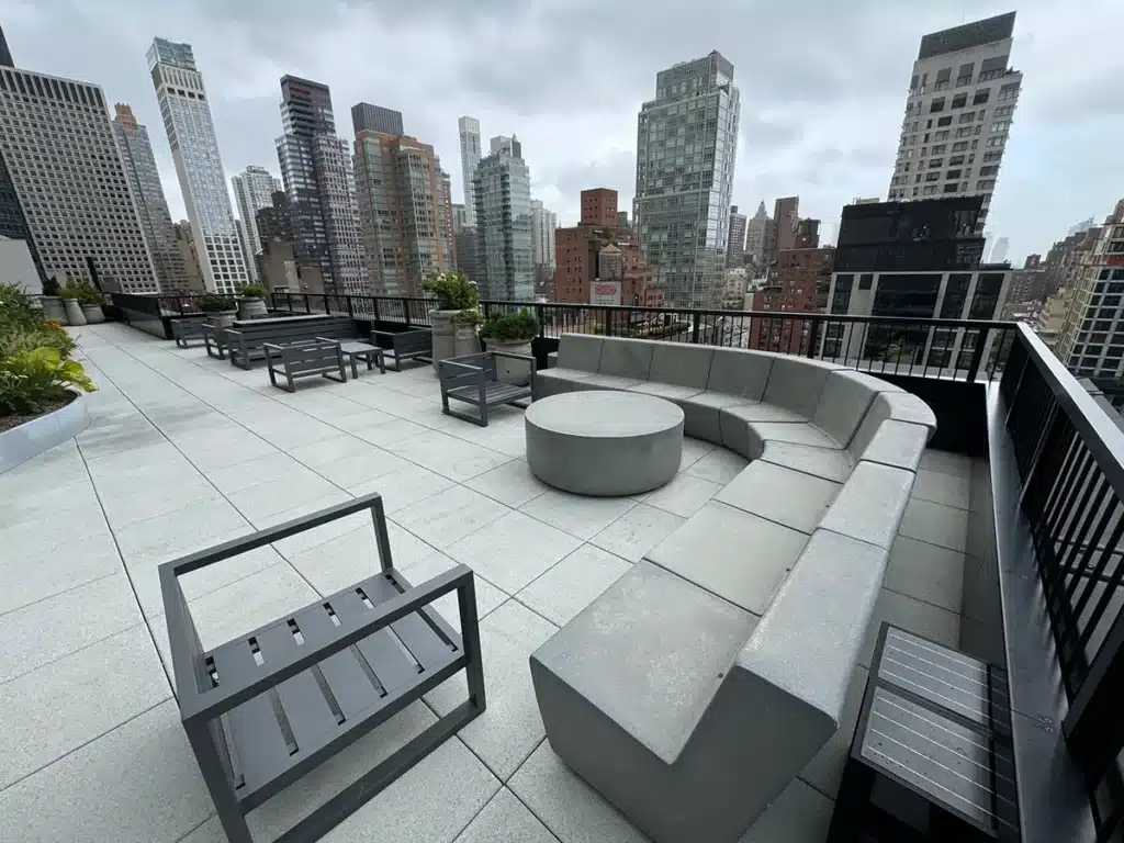 A rooftop patio with modern seating, a circular table, and planter boxes, overlooking a cityscape of tall buildings under a cloudy sky.