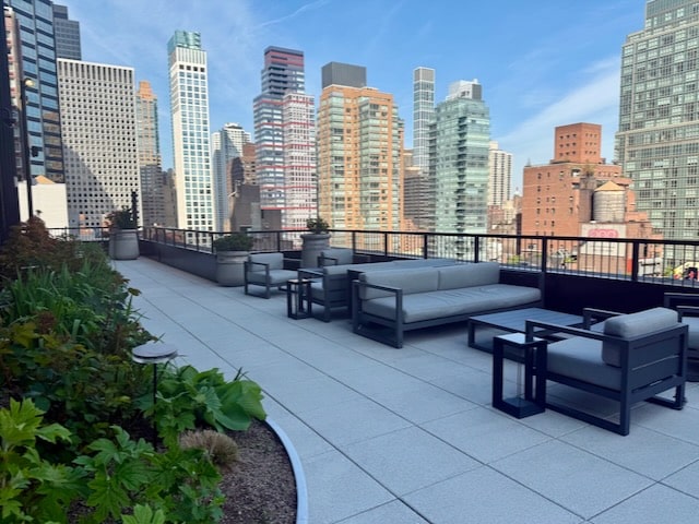Outdoor rooftop lounge with modern seating and planters, overlooking a city skyline with tall buildings under a clear blue sky.
