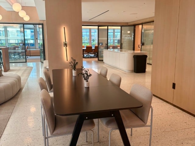 Modern office space with a black table and six beige chairs in the foreground, large windows, light wood walls, and contemporary lighting fixtures.