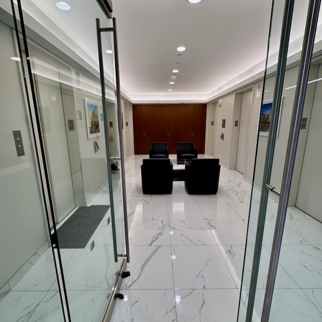 Modern office lobby with glass doors, white marble floors, black chairs, a small table, and wall art, surrounded by elevator doors and wood-paneled walls at the far end.