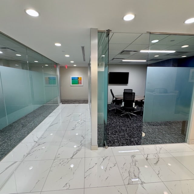 A modern office hallway with glass walls, a tiled floor, and a view into a conference room with a table, chairs, and a wall-mounted TV.