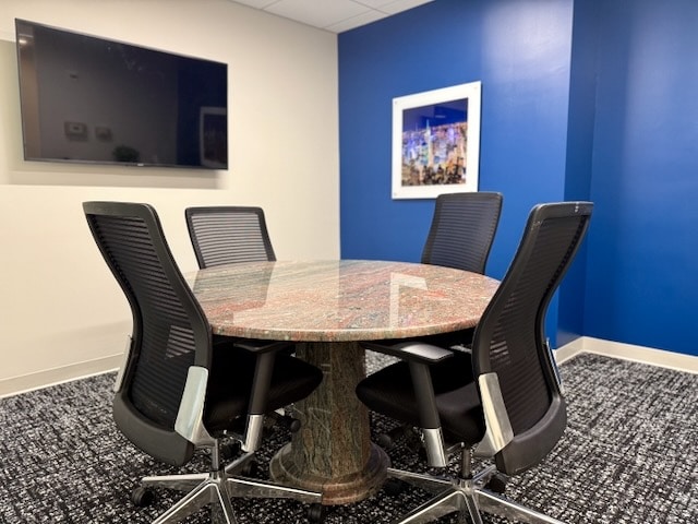 A small conference room with a round marble table, four black office chairs, a wall-mounted TV, and framed artwork on a blue accent wall.