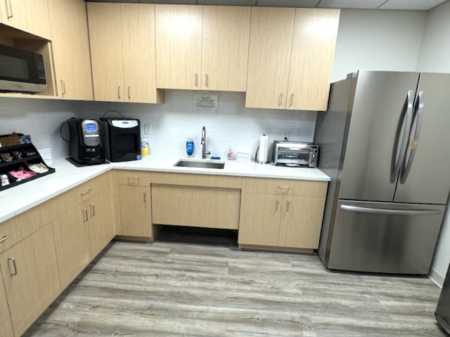 Office kitchen with light wood cabinets, stainless steel refrigerator, microwave, coffee machines, toaster oven, and sink on a wood-patterned floor.