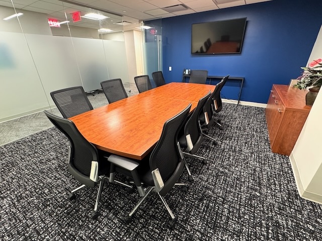 A modern conference room with a wooden table, ten black chairs, a wall-mounted TV, cabinets, and glass walls.