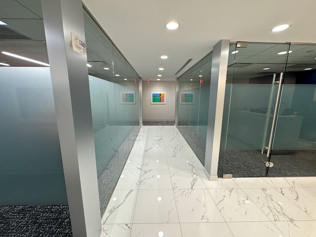 Modern office hallway with glass walls, marble floor tiles, and two colorful abstract artworks hanging at the end of the corridor.