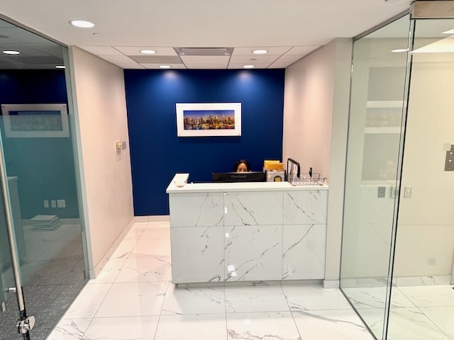 A receptionist sits behind a marble-patterned desk in a modern office lobby with a blue accent wall and framed artwork.