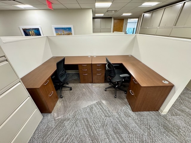 Two empty office workstations with brown desks, black chairs, and white partition walls in a modern office setting.