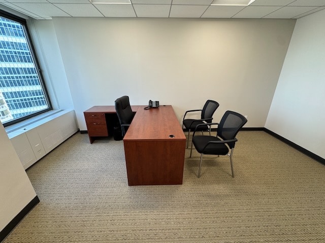 A small office with a wooden desk, two black guest chairs, an office chair, a phone, and a window overlooking tall buildings.