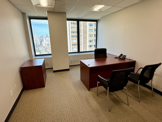 A small office with a wooden desk, two chairs, a side cabinet, a phone, and a window overlooking city buildings.