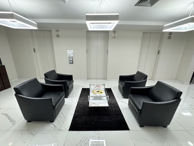 A modern waiting area with four black armchairs around a coffee table with magazines on a black rug, set in front of two closed elevator doors.