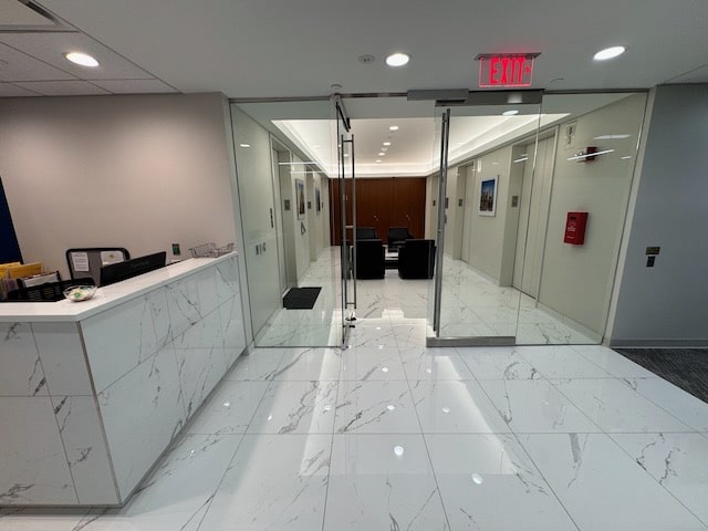 Modern office lobby with marble flooring, glass doors, a reception desk on the left, and seating area visible beyond the doors.