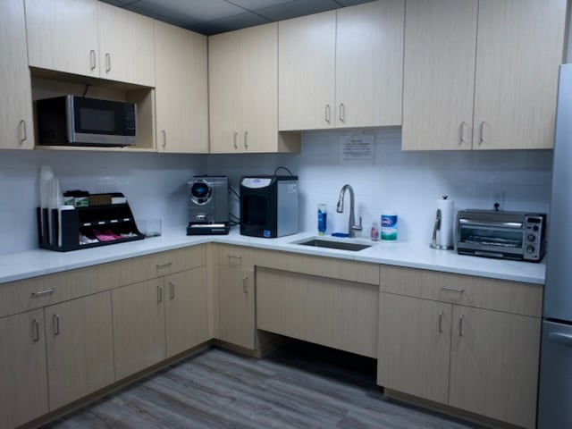 A break room kitchen with light wood cabinets, a sink, microwave, coffee station, toaster oven, and supplies on white countertops.