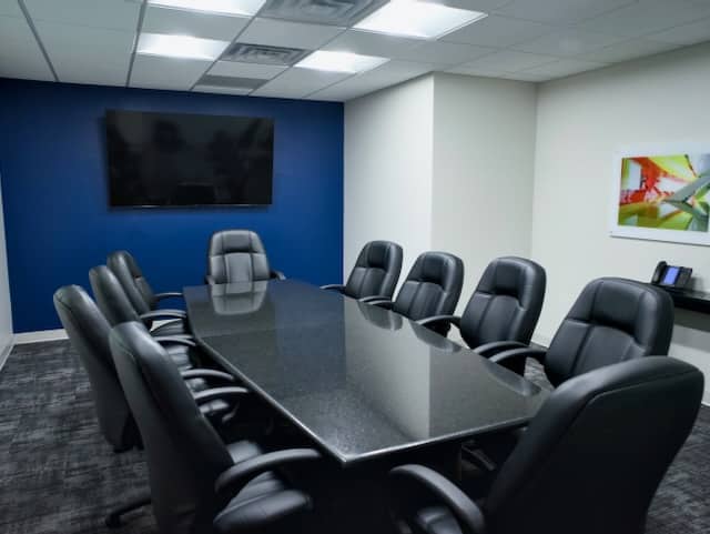 A modern conference room with a long black table, eight black leather chairs, a wall-mounted TV, and a colorful framed picture on the wall.