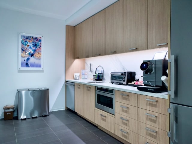 Modern office kitchenette with light wood cabinets, built-in microwave, countertop appliances, a coffee machine, and a double trash bin. Framed artwork hangs on the wall.