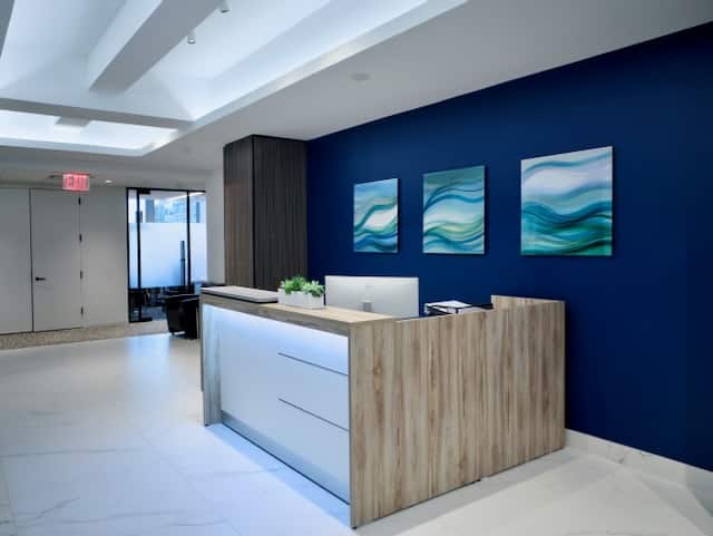 A modern office reception area with a wooden desk, blue accent wall, three abstract paintings, white floors, and potted plants.