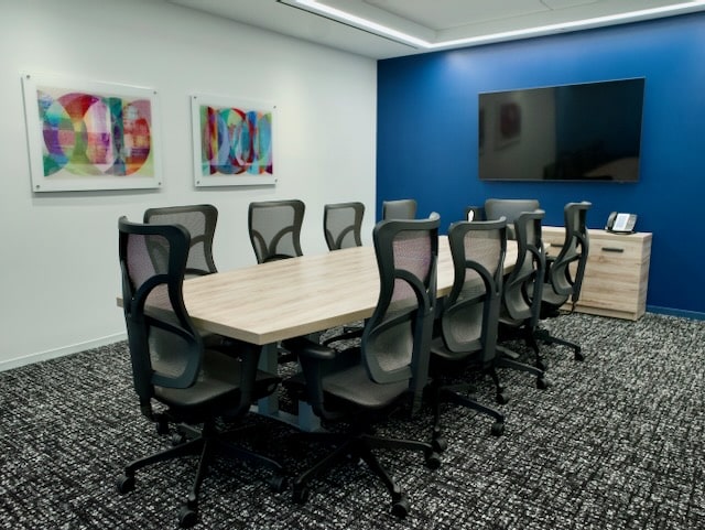 Conference room with a wooden table, ten office chairs, wall-mounted TV, phone on a cabinet, and two abstract colorful artworks on the wall.