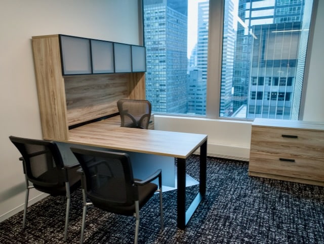 Modern office with a wooden desk, two guest chairs, a storage cabinet, and large windows overlooking tall city buildings.