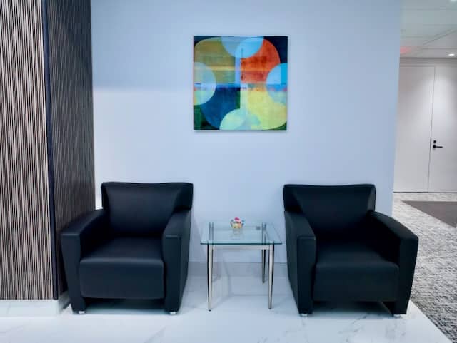 Two black armchairs separated by a glass table with a small bowl of candy, beneath a colorful abstract painting on a white wall.