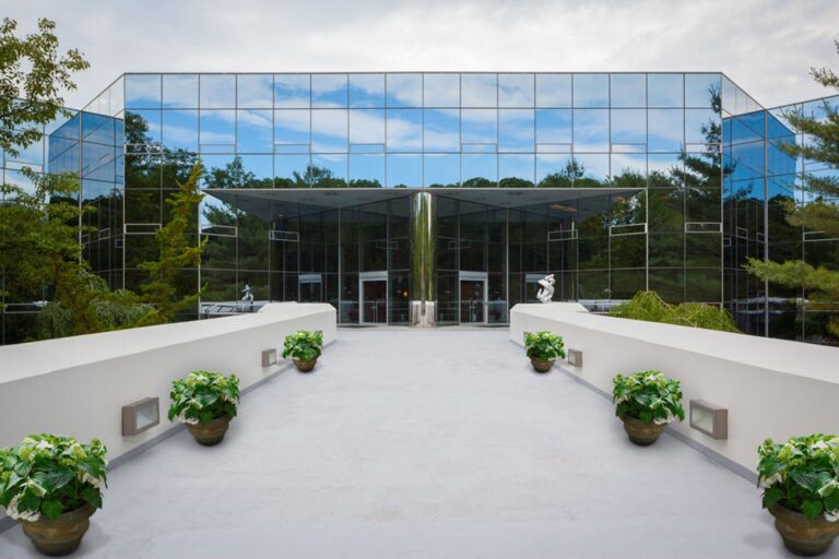 Modern glass building with mirrored windows, viewed from a walkway lined with potted plants.