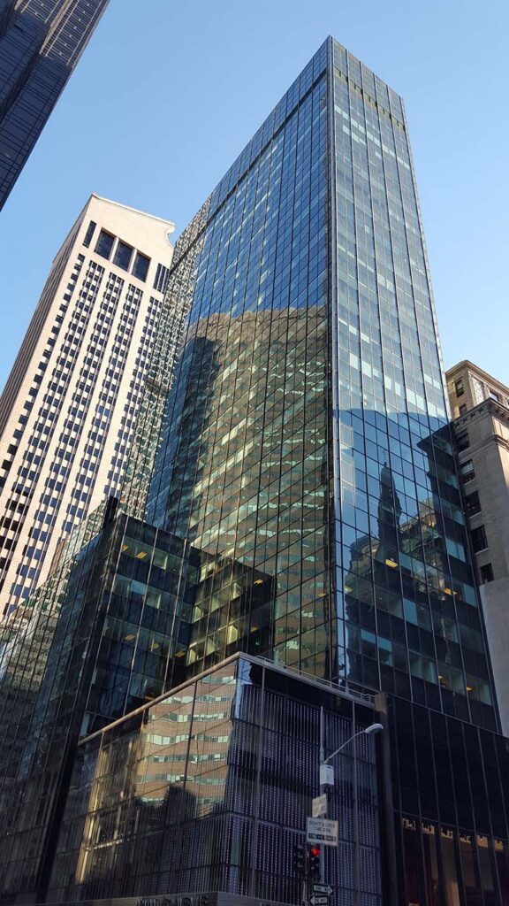 Tall glass skyscrapers reflect nearby buildings under a clear blue sky in a cityscape view.