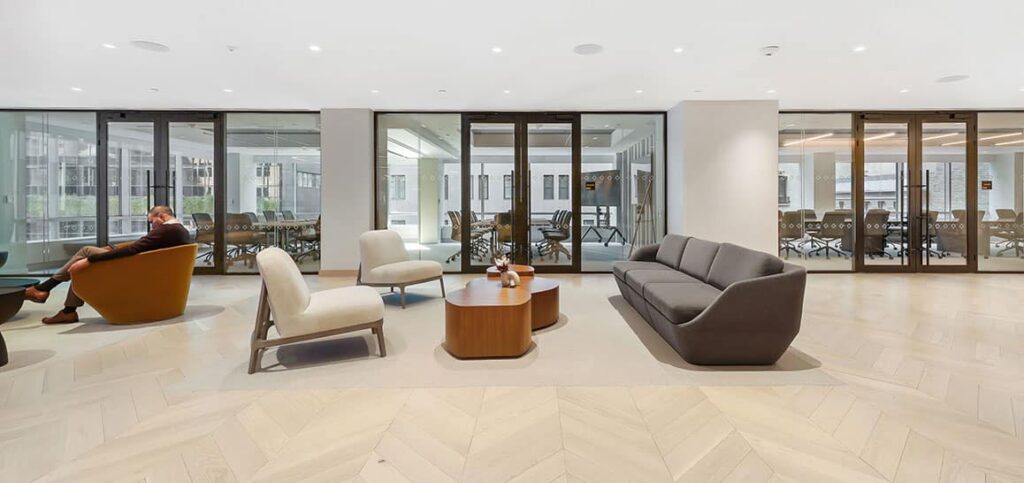 Modern office lounge with armchairs, a sofa, coffee tables, and glass-walled meeting rooms in the background.