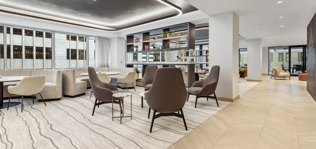 Modern office lounge with cozy chairs, tables, open shelves, large windows, and neutral color tones.