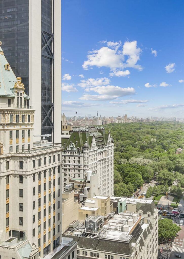 Tall buildings beside Central Park in New York City under a bright blue sky with scattered clouds.