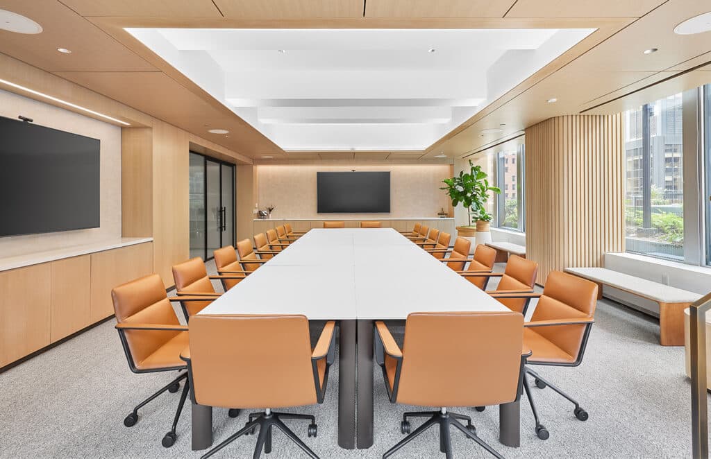 Modern conference room with a large table, tan chairs, two screens, and large windows letting in natural light.