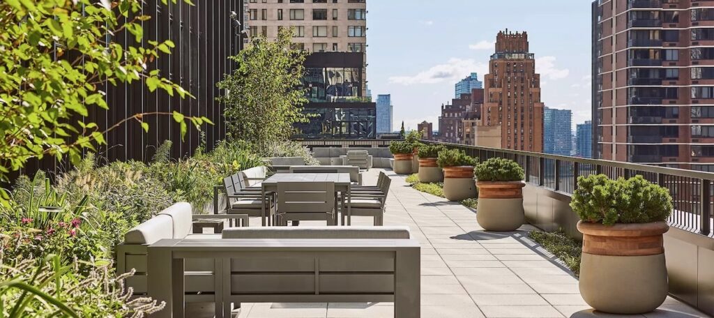 Modern rooftop terrace with seating, potted plants, and city skyscrapers in the background on a sunny day.