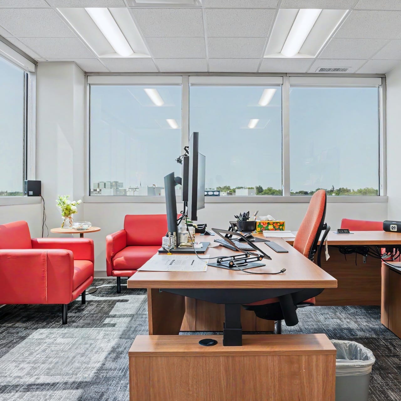 Modern office with two desks, red chairs, computers, large windows, and city view in the background.