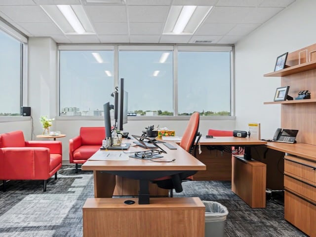 Modern office with wood desk, dual monitors, red chairs, shelves, and large windows with city view.