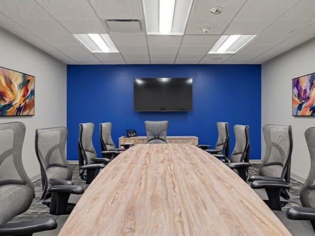 Modern conference room with a long wooden table, mesh chairs, TV on blue wall, and abstract art on walls.