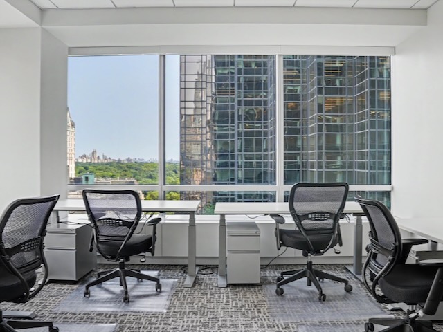 Modern office with empty chairs, desks, and large windows overlooking city buildings and greenery outside.