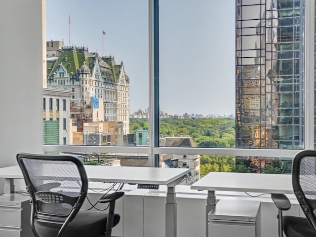 Office desks by a window with a cityscape, historic building, and trees visible outside.
