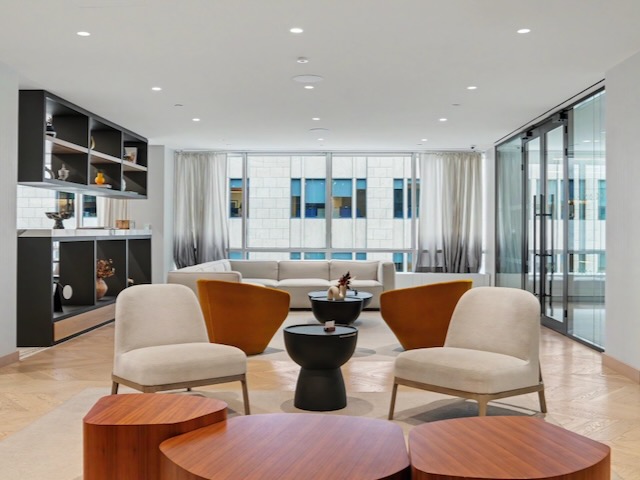 Modern lounge with beige chairs, round tables, shelves, and large windows letting in natural light.