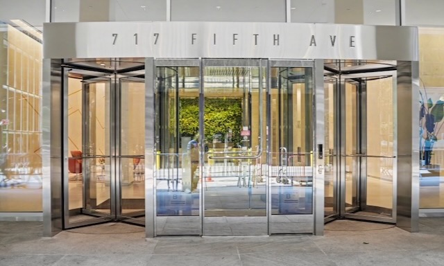 Glass doors and revolving doors at the entrance of 717 Fifth Ave, with reflections and street view, welcome professionals to a world of sophistication—perfect for those seeking Private Furnished Office Suites or an executive office in the heart of Manhattan.