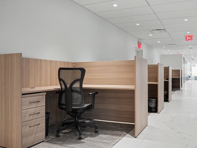 Modern office cubicles with wood panels and mesh chairs in a bright, empty hallway.