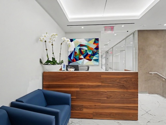 Modern office lobby with wood reception desk, blue chairs, orchids, and colorful abstract art on the wall.