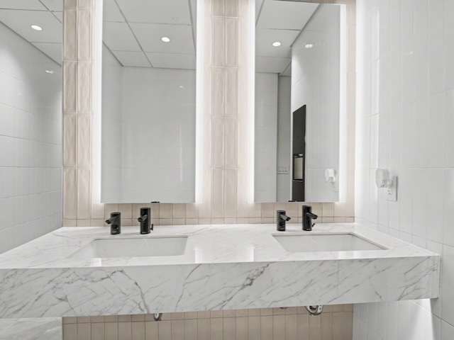 Modern bathroom with a marble double-sink vanity, black faucets, and large backlit mirrors on a tiled wall.