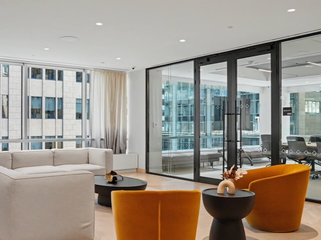 Modern office lounge with white sofa, yellow chairs, black tables, and glass walls overlooking buildings.
