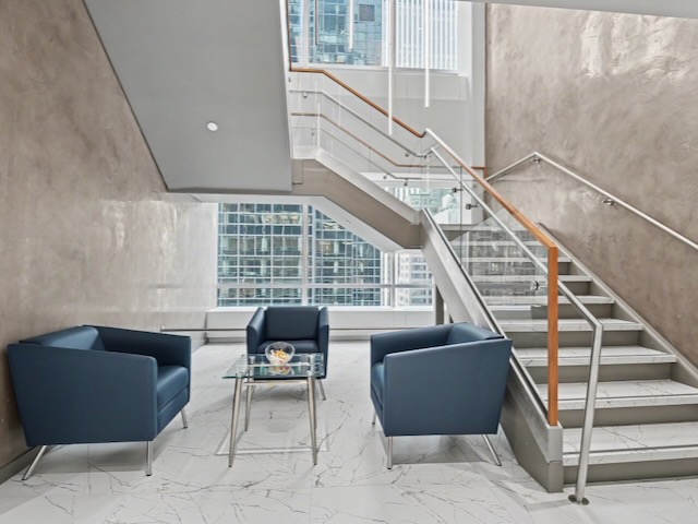 Three blue chairs and a glass table sit by a staircase in a modern, bright lobby with large windows.