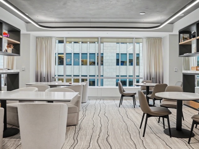 Modern office lounge with round tables, beige chairs, large windows, and city building views outside.